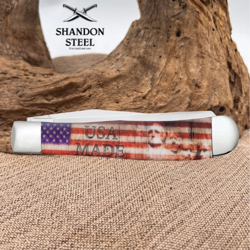 A folding pocket knife with a distressed American flag graphic and 'USA MADE' text, resting on a burlap surface in front of a piece of driftwood. The brand logo 'SHANDON STEEL' is visible in the upper left.