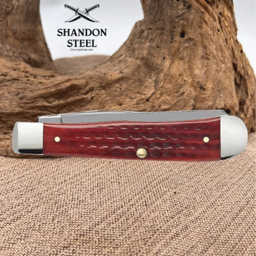 A folding pocket knife with a reddish-brown textured handle and silver-toned metal bolsters, displayed on a burlap surface with a piece of driftwood in the background. The brand logo 'SHANDON STEEL' is visible in the upper left.