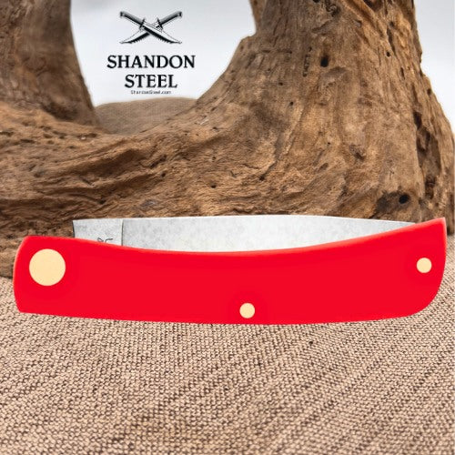 A red folding pocket knife with gold-toned brass pins and a silver blade, displayed on a burlap surface with a piece of driftwood and a white Shandon Steel logo card in the background.