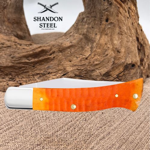 A folding pocket knife with a bright orange textured handle and a silver blade, displayed on a burlap surface with a large piece of driftwood in the background. The brand logo 'SHANDON STEEL' is visible in the upper left.