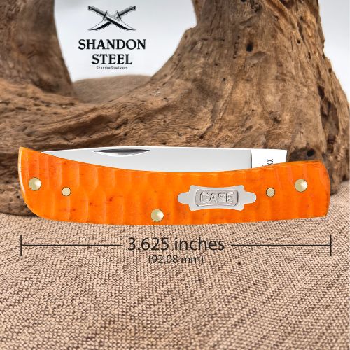 An orange textured folding knife with a silver blade, displayed on a burlap surface with a piece of driftwood in the background. The brand logo 'Shandon Steel' is visible in the top left, and a dimension overlay indicates the knife is 3.