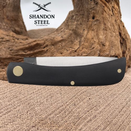 A black folding pocket knife with brass hardware lies open on a burlap surface in front of a large piece of driftwood. The Shannon Steel logo is visible in the upper left.