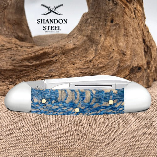 A pocket knife with a blue and beige patterned handle and silver-toned steel blade, displayed on a burlap surface in front of a large piece of driftwood. The Shandon Steel logo is visible in the upper left.