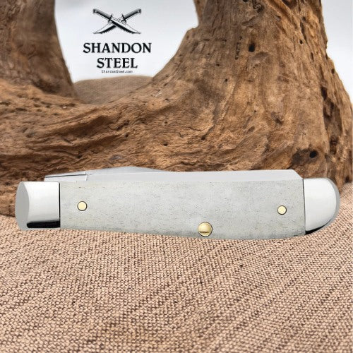 A folding pocket knife with a textured light gray handle and brass-toned pins, displayed on a burlap surface with a piece of driftwood and a white background featuring the 'Shandon Steel' logo.