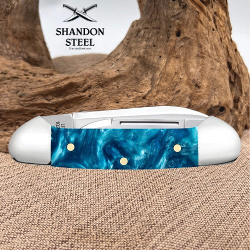 A folding pocket knife with a white handle and a blue marbled inlay, displayed on a burlap surface with a piece of driftwood in the background. The brand logo 'SHANDON STEEL' is visible in the upper background.