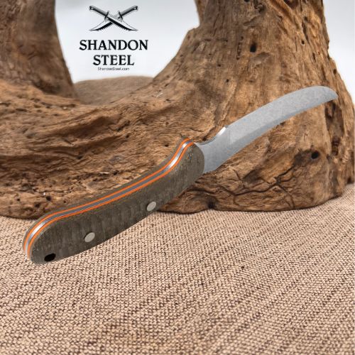 A folding pocket knife with a curved silver blade and a textured gray handle accented by orange and red stripes, displayed on a piece of driftwood over a burlap surface. The Shandon Steel logo is visible in the upper left.