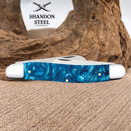 A folding pocket knife with a blue marbled resin handle and silver metal blades, displayed on a burlap surface in front of a piece of driftwood. The brand logo 'SHANDON STEEL' is visible in the upper background.