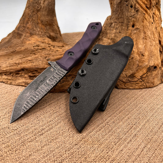 A handmade knife with a purple handle and a patterned steel blade, accompanied by a black Kydex sheath, displayed on a woven mat next to a wooden object.