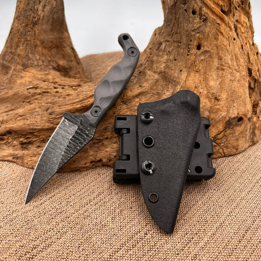 A small, compact folding knife with a grey blade and black handle, placed next to its black Kydex sheath on a textured background.