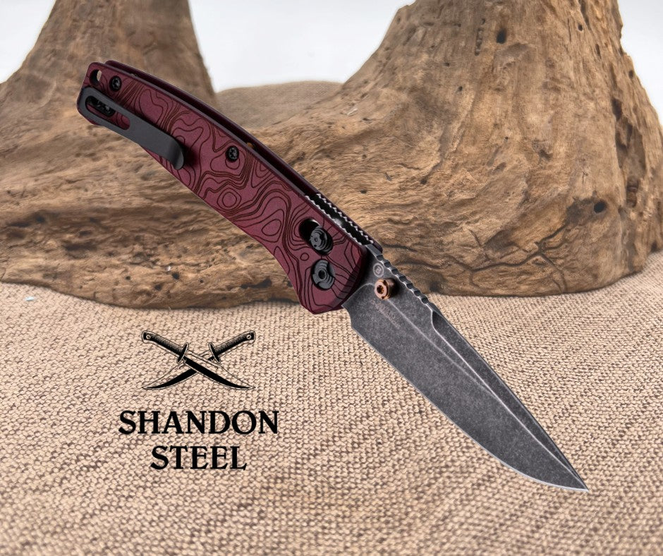 Folding knife with maroon handle and black blade on a textured surface, Shandon Steel logo visible.