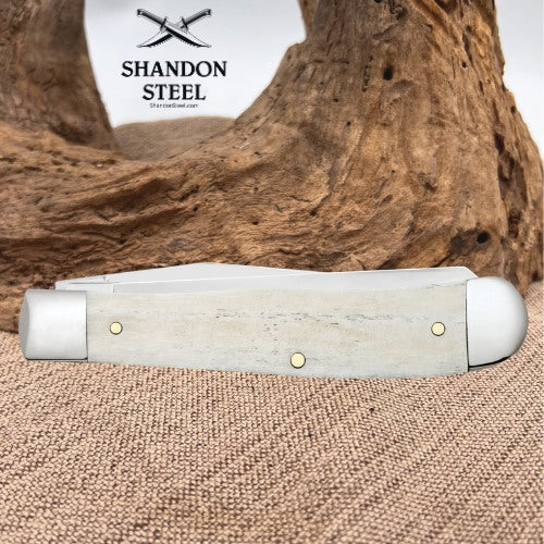 A folding pocket knife with a light gray bone or horn handle and brass pins, displayed on a burlap surface with a piece of driftwood in the background. The brand logo 'SHANDON STEEL' is visible in the upper left.