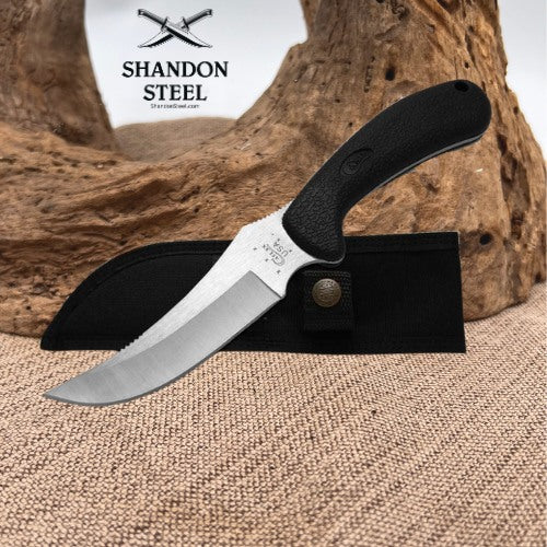 A fixed-blade knife with a black textured handle and a silver blade, resting on a black sheath. The background features a rustic wood log and burlap fabric, with the 'Shandon Steel' brand logo visible in the upper left.