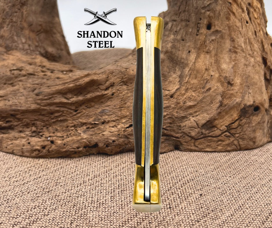 A folding pocket knife with a gold-toned steel bolster and black and gold handle scales, standing upright on a burlap surface with a piece of driftwood in the background. The brand logo 'SHANDON STEEL' is visible in the upper left.