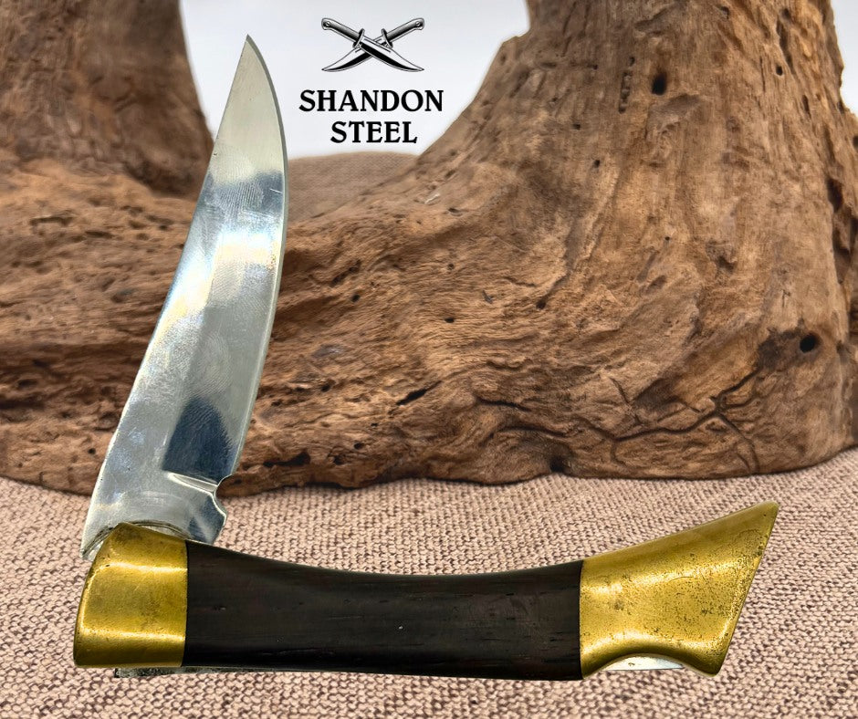 A folding pocket knife with a polished steel blade, a black handle, and brass end caps, displayed against a rustic wood and burlap backdrop. The brand logo 'Shandon Steel' is visible at the top.