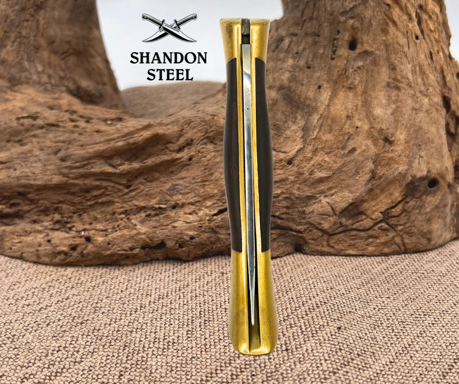 A vertical close-up of a folding knife handle with a polished gold-toned metal clip and dark textured grip panels, standing upright in front of a rustic wood prop on a burlap surface. The brand name 'SHANDON STEEL' with crossed knives logo is visible in the upper left.
