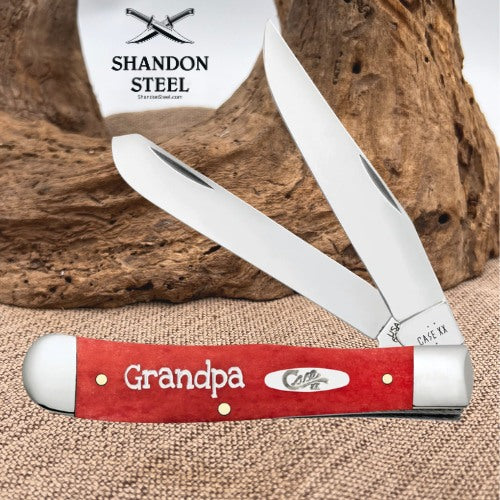 A folding pocket knife with a red handle engraved with 'Grandpa' and 'Case Co.' is displayed in front of a rustic wood background.