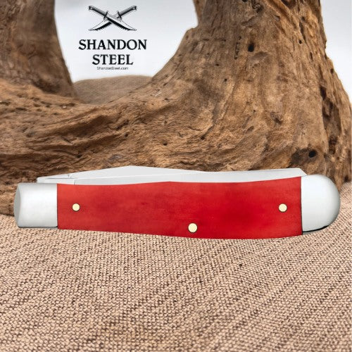 A red and white pocket knife with brass pins rests on a burlap surface in front of a large piece of driftwood. The brand logo 'SHANDON STEEL' is visible in the upper left.