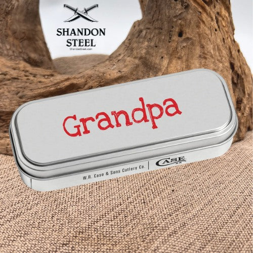 A silver metal tin engraved with the word 'Grandpa' in red, placed on a burlap surface with driftwood in the background. The Shandon Steel logo is visible in the upper left.