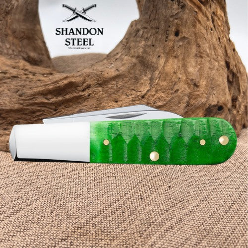 A folding pocket knife with a bright green textured handle and a white bolster, resting on a burlap surface with a piece of driftwood in the background. The 'SHANDON STEEL' brand logo is visible in the upper left.