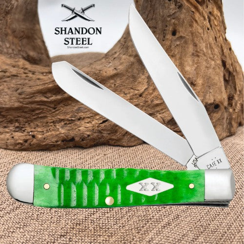 A green and silver folding pocket knife with two open blades, displayed on a burlap surface in front of a rustic wood log. The brand logo 'SHANDON STEEL' is visible on a white tag behind the knife.