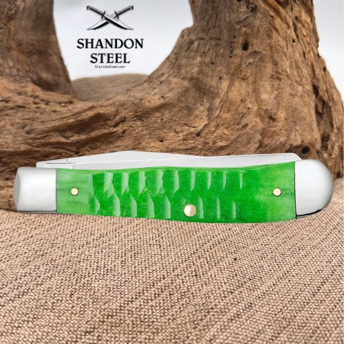 A folding pocket knife with a bright green textured handle and white metal bolsters, resting on a burlap surface in front of a piece of driftwood. The brand logo 'SHANDON STEEL' is visible in the upper left.