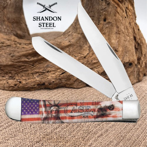 A folding pocket knife with a printed American flag and Statue of Liberty handle, shown open with a wood-grain background and a white brand tag reading SHANDON STEEL.
