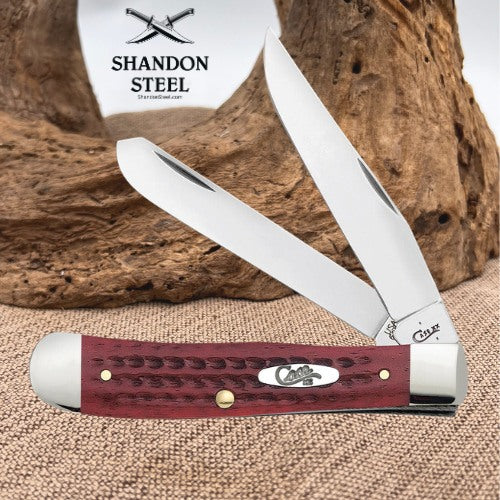 A folding pocket knife with a reddish-brown textured handle and silver stainless steel blades, displayed on burlap in front of a wood log. The background features a white card with the 'Shandon Steel' logo.