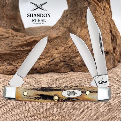 Two folding pocket knives with cream and black bone handles are displayed against a rustic wood driftwood background on a burlap surface. The left knife is closed, and the right knife is open, showing a silver steel blade and a black polished edge.