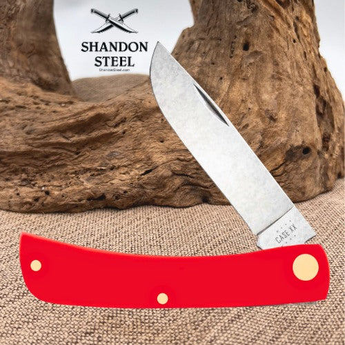 A folding pocket knife with a bright red handle and a satin-finished silver blade, displayed against a rustic wood driftwood and burlap background. The brand logo 'SHANDON STEEL' is visible in the upper left.