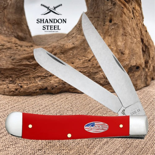 A red and silver folding pocket knife with a small American flag emblem on the handle, displayed in front of a rustic wood background with a Shandon Steel logo card visible behind it.