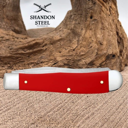 A red folding pocket knife with silver metal bolsters and brass pins, displayed on a burlap surface with a large piece of driftwood in the background. The Shandon Steel logo is visible in the upper left corner.