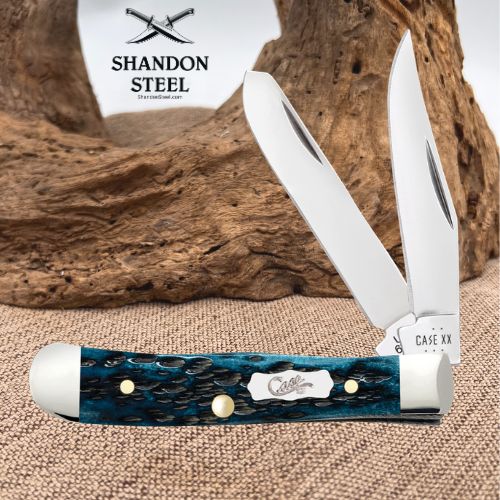 A folding pocket knife with a blue marbled handle and silver blades, displayed on a burlap surface with a piece of driftwood in the background. The brand logo 'SHANDON STEEL' is visible in the upper left.