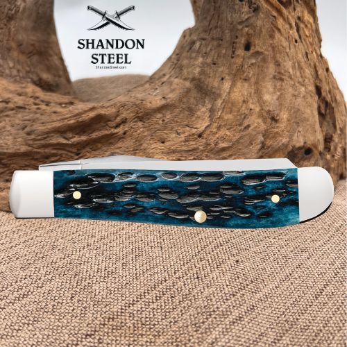 A folding pocket knife with a textured blue and black inlay handle and white end caps, displayed on a burlap surface with a piece of driftwood in the background. The brand logo 'SHANDON STEEL' is visible in the upper left.