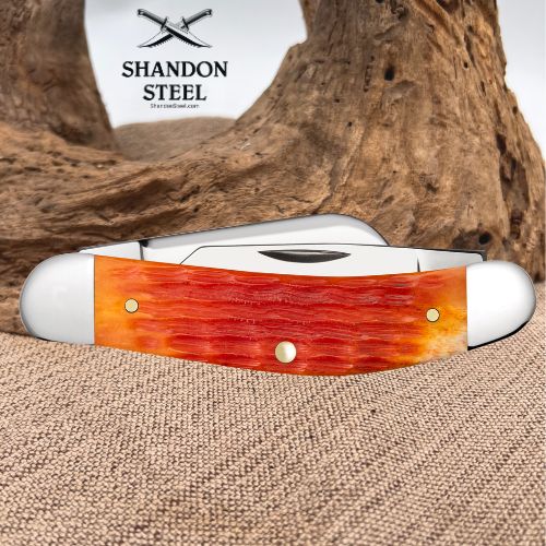 A folding pocket knife with a textured orange and red bone handle and silver-toned metal blades, displayed on a burlap surface with a piece of driftwood in the background. The brand logo 'SHANDON STEEL' is visible in the upper left.