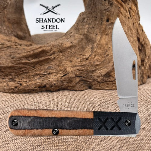 A folding pocket knife with a silver blade and a handle made of tan wood and black leather, displayed in front of a piece of driftwood on a burlap surface. The Shandon Steel logo is visible in the upper left, and 'CASE XX' is stamped on the blade.