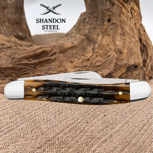 A folding pocket knife with a brown and black patterned handle and silver metal blades, displayed on a burlap surface with a piece of driftwood in the background. The brand logo 'SHANDON STEEL' is visible in the upper left.