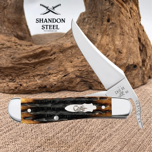 A custom engraved pocket knife with a polished silver blade and a carved handle made of black and brown tortoiseshell, displayed on a burlap surface with a piece of driftwood and a Shandon Steel logo card in the background.