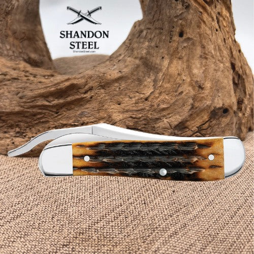 A folding pocket knife with a polished steel blade and handle, featuring a natural brown and black hornback scale inlay, displayed on a burlap surface with a piece of driftwood in the background. The 'SHANDON STEEL' brand logo is visible in the upper left.