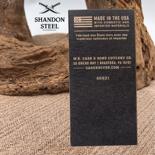 A black metal nameplate or plaque with gold engraved text and an American flag graphic, standing upright on a burlap surface with driftwood in the background. The top left shows a white logo reading SHANDON STEEL.