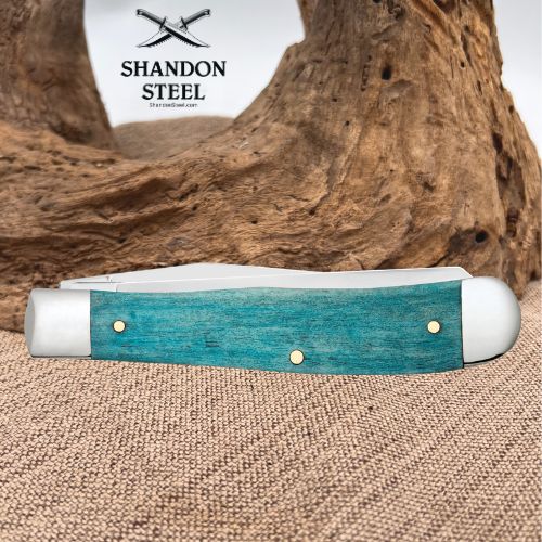 A folding pocket knife with a turquoise blue textured handle and white end caps, displayed on a burlap surface with a piece of driftwood in the background. The brand logo 'SHANDON STEEL' is visible in the upper left.