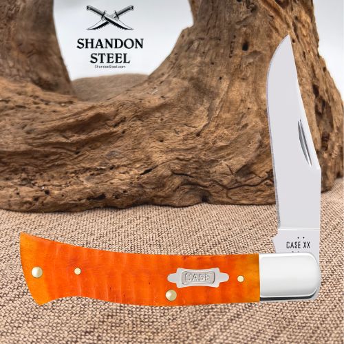 A folding pocket knife with a bright orange textured handle and a silver blade, displayed horizontally in front of a rustic piece of driftwood. The brand logo 'SHANDON STEEL' is visible in the upper left, and the text 'CASE XX' is printed on the bolster of the knife.