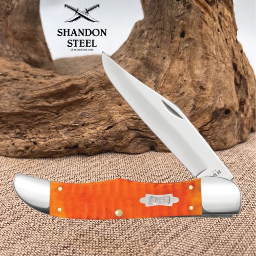 A folding pocket knife with a bright orange textured handle and a silver blade, displayed against a rustic wood driftwood and burlap backdrop. The brand logo 'SHANDON STEEL' is visible in the upper left.