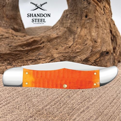 A folding pocket knife with a bright orange textured handle and silver-toned steel blades, displayed on a burlap surface with a large piece of driftwood in the background. The brand logo 'SHANDON STEEL' is visible in the upper left.