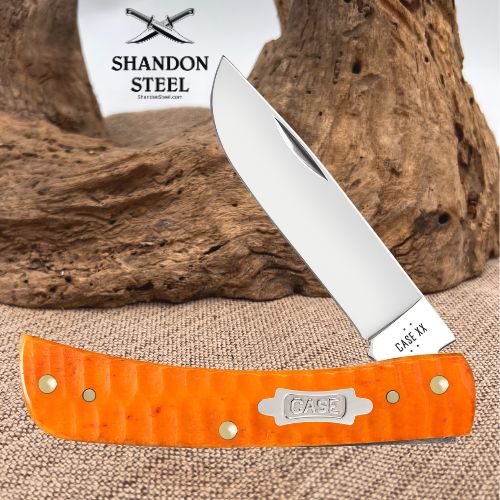 A folding pocket knife with a silver stainless steel blade and an orange textured handle, displayed diagonally in front of a rustic wood log. The brand logo 'SHANDON STEEL' is visible in the upper left, and the 'CASE' logo is engraved on the metal tang and handle badge.
