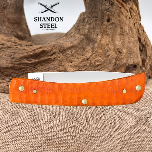 A folding pocket knife with a bright orange textured handle and a silver blade, displayed on a burlap surface with a piece of driftwood in the background. The brand logo 'SHANDON STEEL' is visible in the upper left corner.