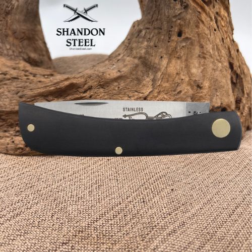 A folding pocket knife with a black handle and stainless steel blade, displayed horizontally on a textured burlap surface with a piece of driftwood in the background. The brand logo 'SHANDON STEEL' is visible in the upper left corner.