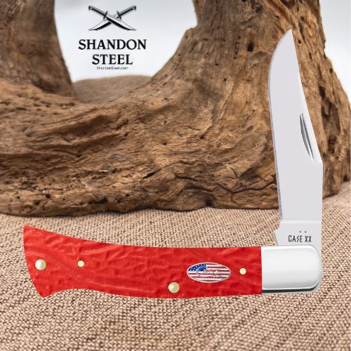 A red leather folding knife with a white metal bolster and blade, displayed horizontally in front of a large piece of driftwood on burlap. The brand logo 'SHANDON STEEL' is visible in the upper left, and 'CASE XX' is printed on the knife blade.