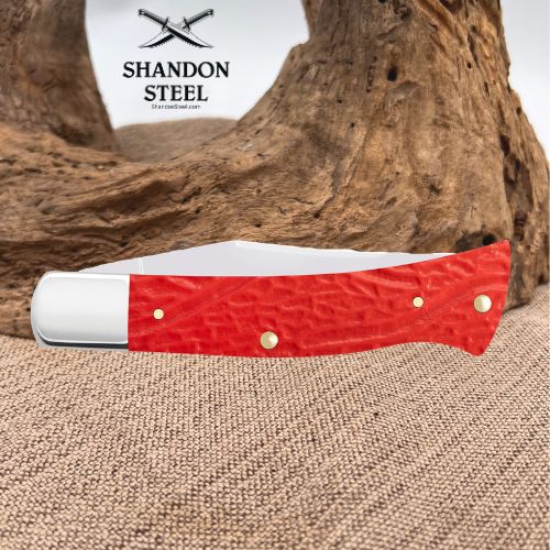 A folding pocket knife with a textured red handle and white bolster, displayed on a burlap surface with a piece of driftwood in the background. The brand logo 'SHANDON STEEL' is visible in the upper left.