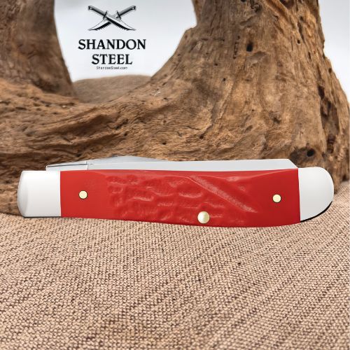 A red and white folding pocket knife with brass pins, displayed on a burlap surface with a piece of driftwood and a white background featuring the 'Shandon Steel' logo.