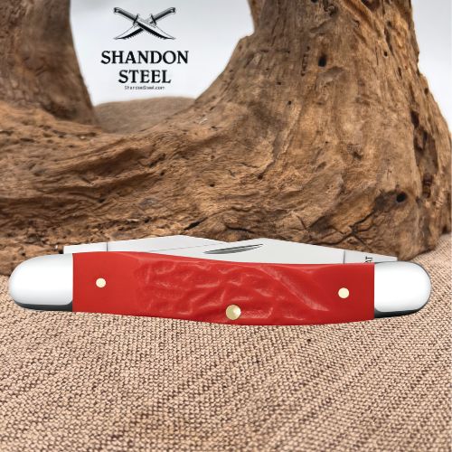A red and white pocket knife with brass rivets, displayed on a burlap surface with a large piece of driftwood in the background. The brand logo 'SHANDON STEEL' is visible in the upper left.